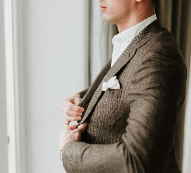 Groom in a brown suit with white shirt and pocket square looking out of the window | Anne-Sophie Benoit Photography