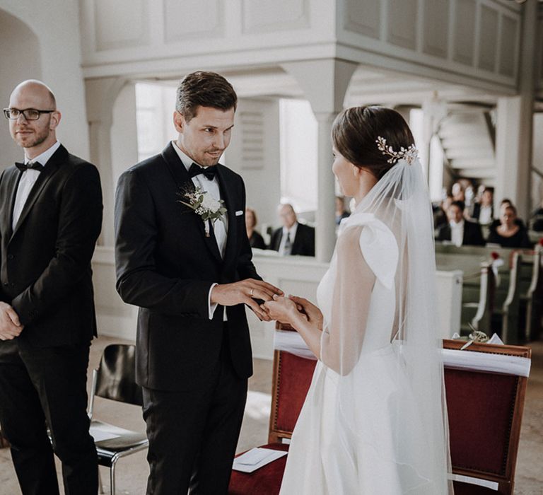 Groom in black Tiger of Sweden tuxedo exchanges rings with Bride in white Jesus Piero Wedding gown and cathedral veil during German wedding ceremony