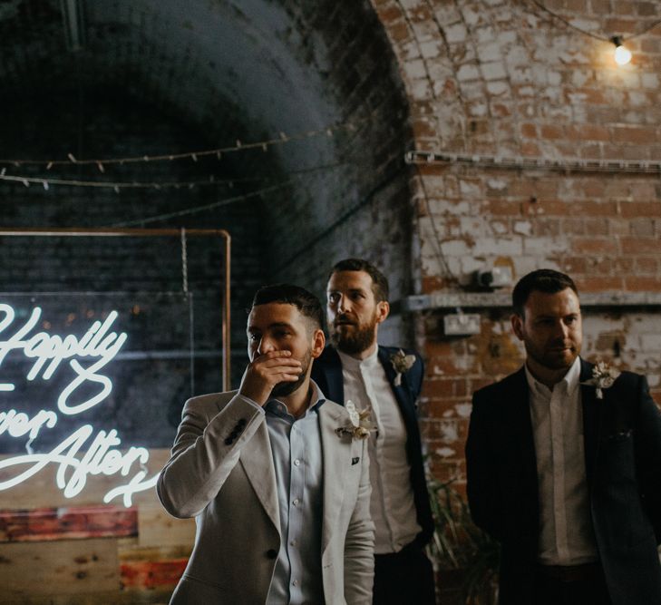 Groom looking shocked with hand over mouth at top of aisle with groomsmen in front of 'happily ever after' neon sign