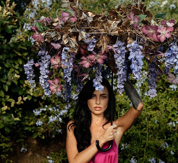 A bridesmaid in a pink jumpsuit holds an umbrella made form flowers. Photography by Stephanie Shenton. 