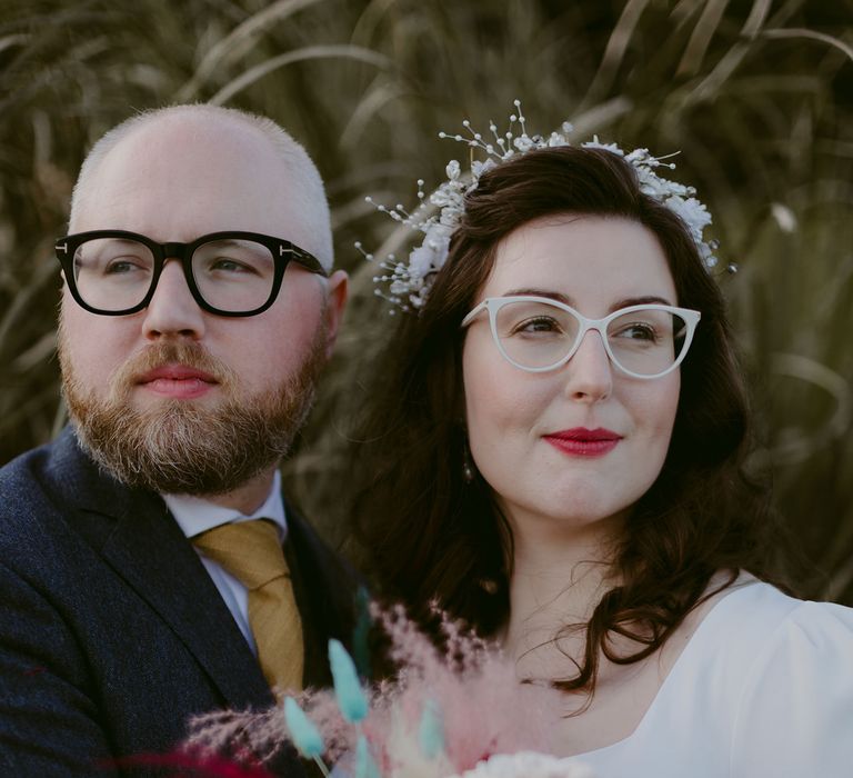 Groom in navy suit and yellow tie stands with bride in white cat eye glasses and bridal headband holding white and pink rose and pampas grass bouquet