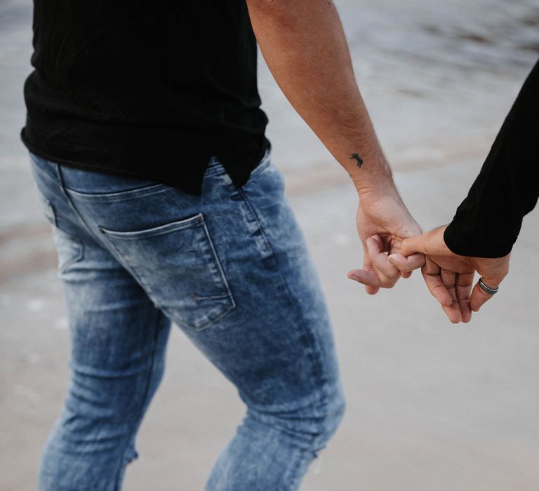 Groom wears light denim jeans and walks barefoot along the sand whilst holding hands