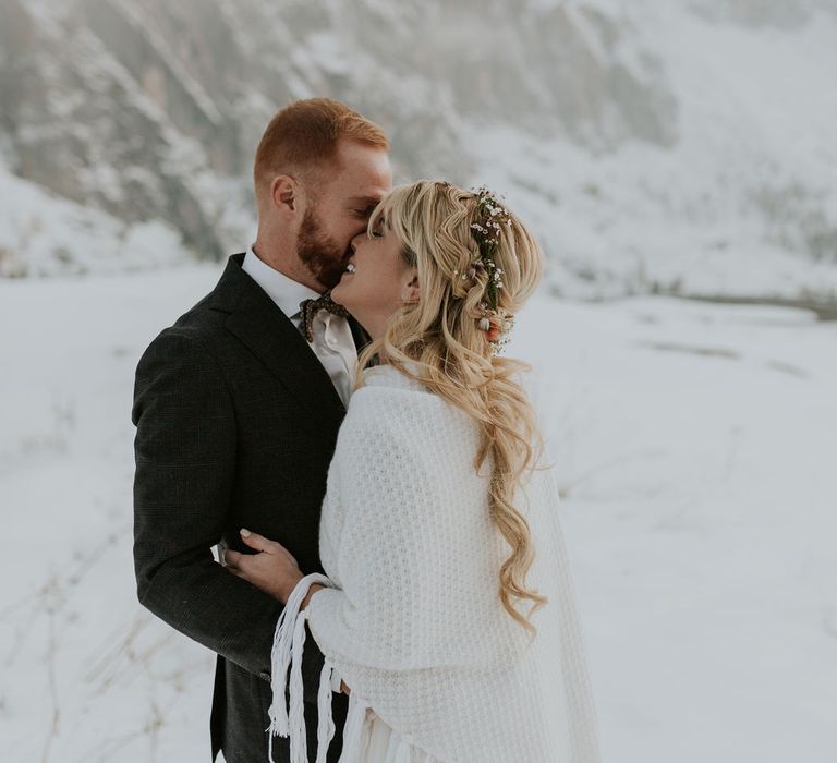 Groom in a dark suit and bow tie kissing his bride in a shawl and flower crown at their Dolomites wedding 