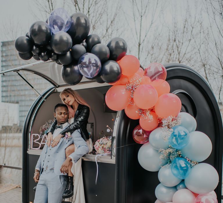 Bride and groom standing in front of their bar van with a pastel balloon installation, neon bar sign and spray painted gypsophila clouds 