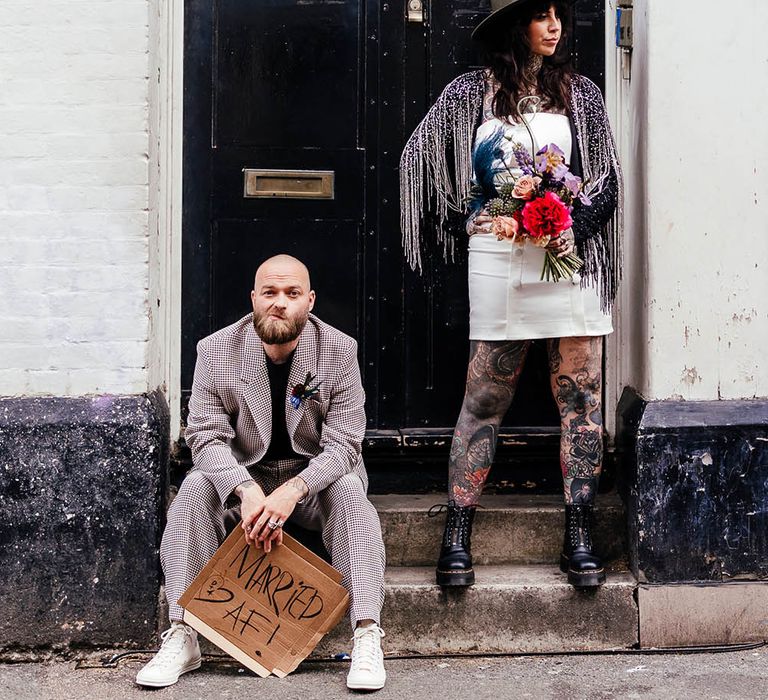 Alternative bride and groom standing in a doorway with groom in a check suit and bride in tassel jacket and hat 