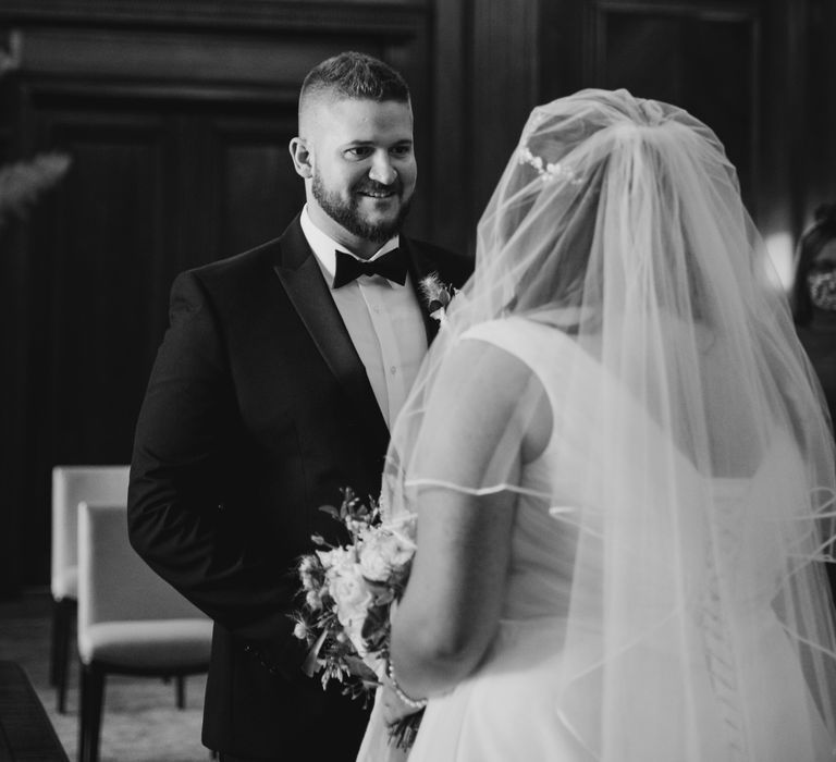 Black & white image of groom wearing tuxedo during wedding ceremony looking at his bride