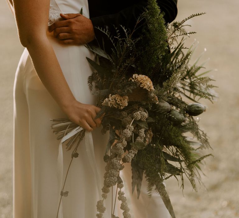 Bride holding a large tropical green wedding bouquet with ferns, dried poppy heads and celosias