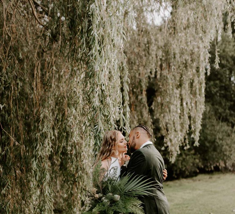 Bride and groom standing under a willow tree, holding a large green tropical bouquet