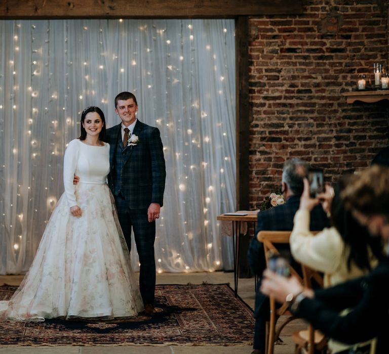 Bride and groom standing at the altar embracing with a fairy light and chiffon backdrop 