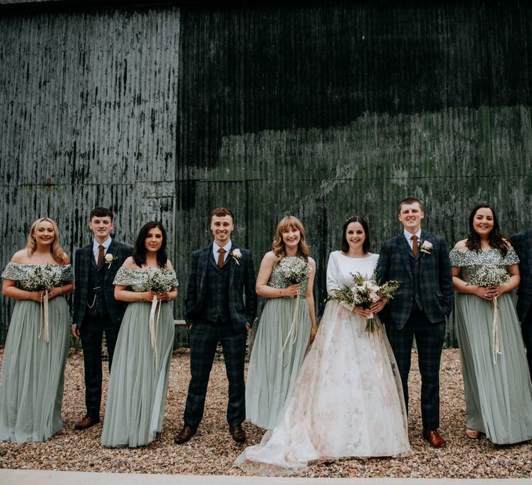 Wedding party portrait with bridesmaids in green chiffon and sequin dresses, groomsmen in navy check suits and the bride in a floral skirt wedding dress 