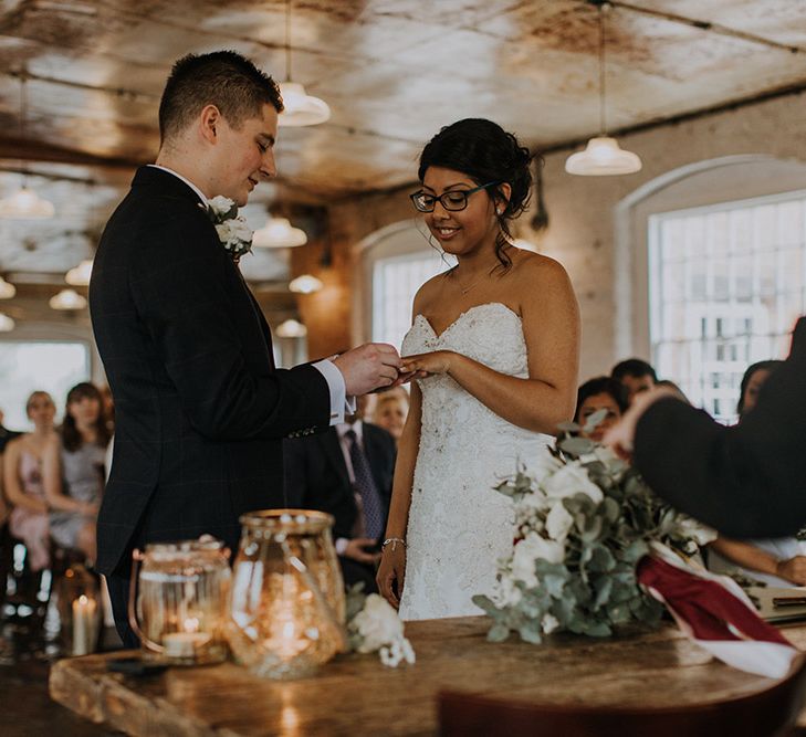 An Asian bride in a Sophia Tolli gown and glasses holds her hand out as her new husband places a wedding ring on her finger. 