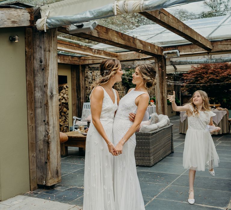 Bride holds wife on their wedding day leaning in to kiss as daughter throws confetti