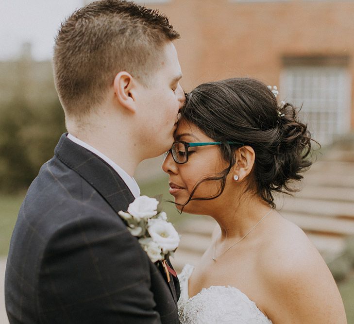 A groom kisses a bride on the forehead. She wears Sophia Tolli and black rimmed glasses.