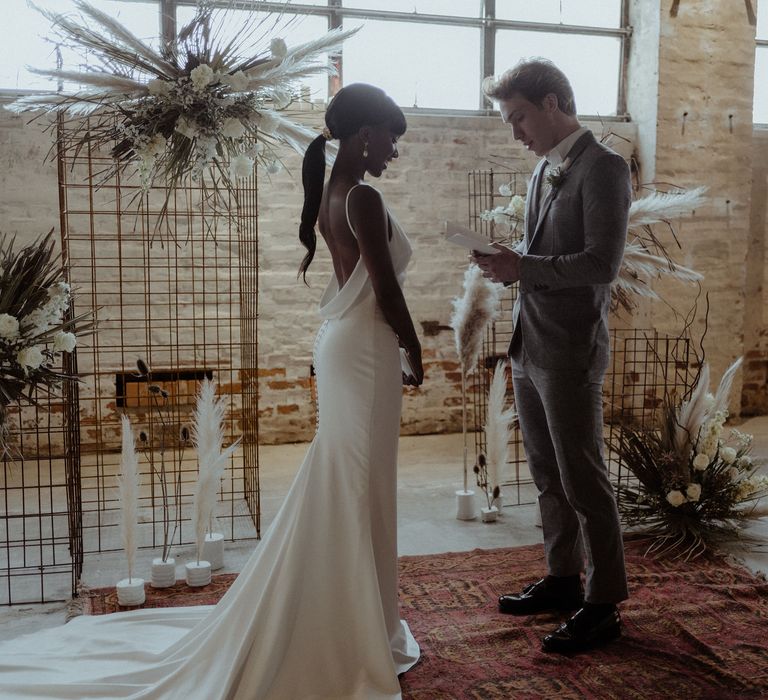 A bride and groom stand in front of a metal frame structure and he reads his vows from a book.