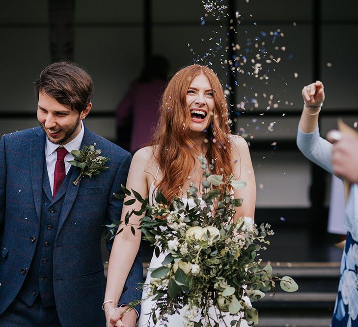 Confetti moment with groom in a navy and red check suit and bride in a slip wedding dress holding an oversized green and white bouquet 