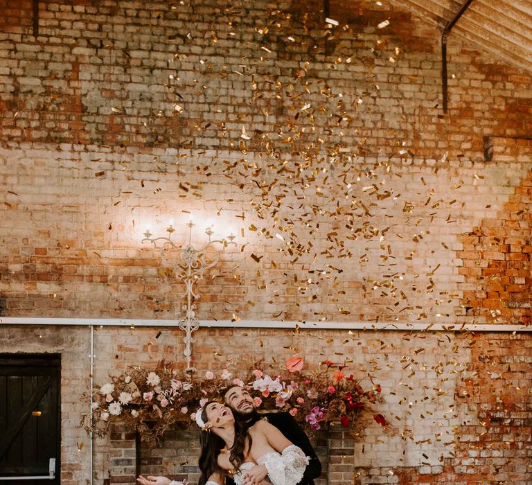 Groom in a black suit embracing his bride in a strapless appliqué gown with detachable sleeves under an explosion of gold confetti 