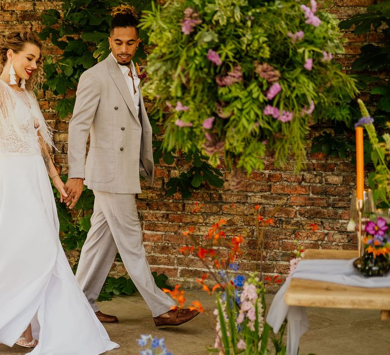 Groom in a beige double breasted jacket holding hands with his bride in bridal separates with tassel coverup