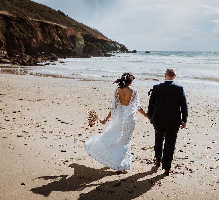 Bride & groom walk along beach whilst holding hands as the wind blows brides lace wedding gown 