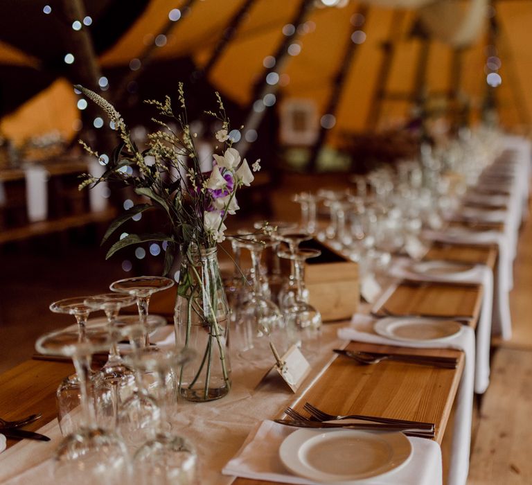 Rustic wooden wedding breakfast table with white table runner, wild flowers in glass bottles and wooden benches inside tipi wedding reception at home farm wedding