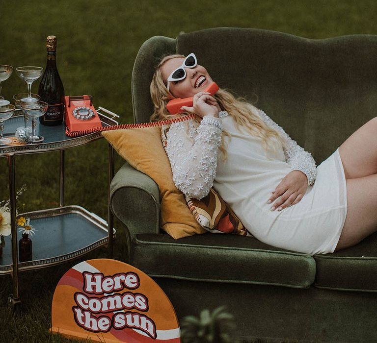 Bride in a short wedding dress laying on a retro sofa answer a telephone with sunglasses on