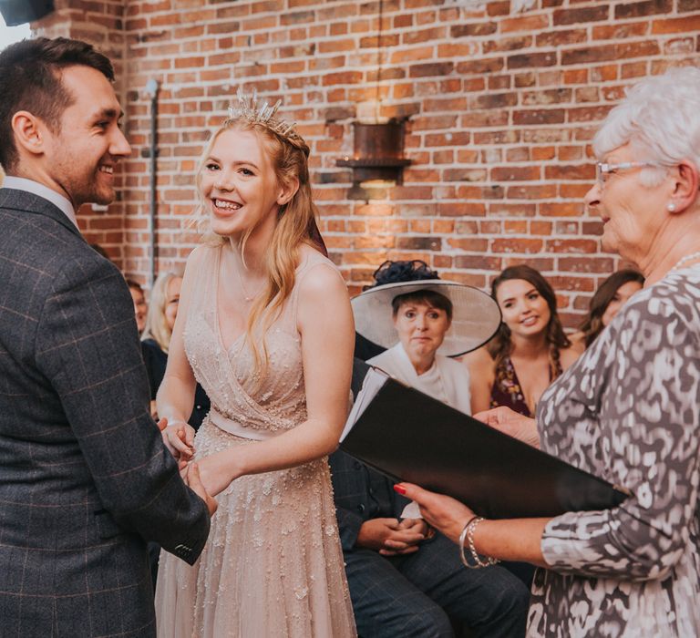 Bride in pearl wedding dress and groom in checked suit smile whilst holding hands during barn wedding ceremony