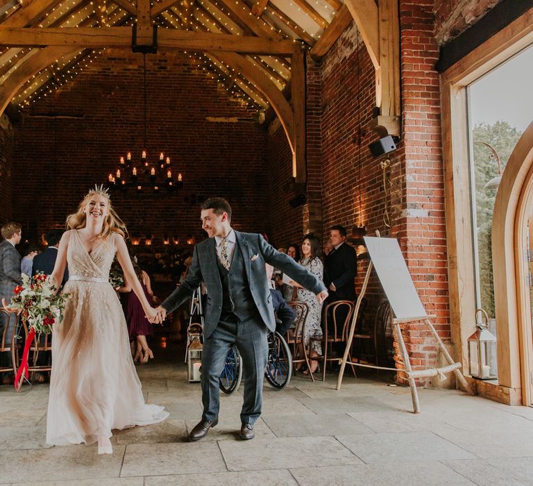 Bride in pearl wedding dress holding red floral bouquet and groom in checked suit hold hands inside barn after wedding ceremony