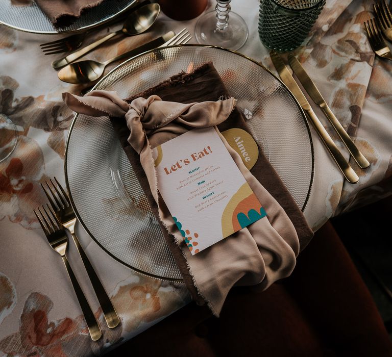 Place setting with gold rimmed charger plate, gold cutlery and retro 70s wedding stationery 