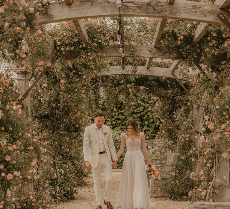 Bride in a sparkly Made With Love dress holding hands with her groom in a stone coloured suit as they walk under the pergola at Euridge Manor 