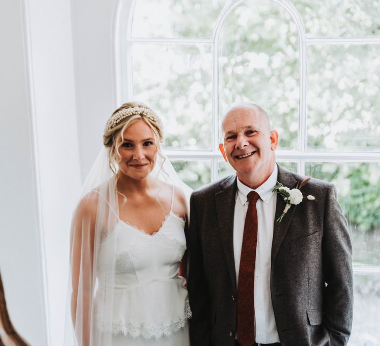 Bride stands with her father on her wedding day in front of window as they prepare to walk down the aisle