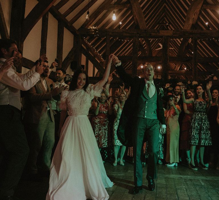 Bride in white lace puffed sleeve Daalarna wedding dress holds arm up with groom in morning coat and waistcoat as they dance in barn surrounded by wedding guests at Loseley Park in Surrey