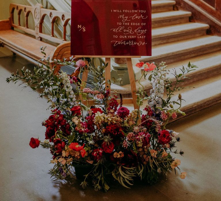 Burgundy acrylic wedding quote sign on a wooden easel with red, purple and pink floral arrangement decor at the bottom of the stairs at Dulwich College 