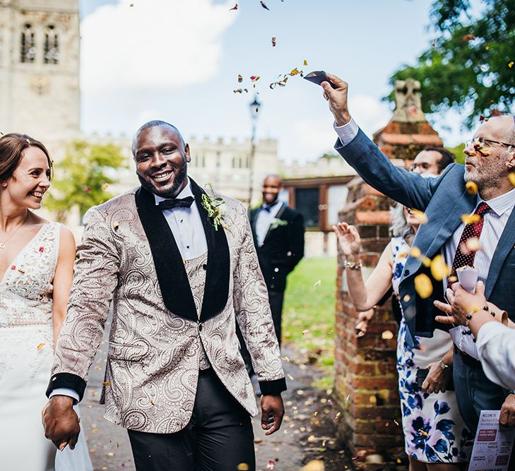 A newley married couple walk through a shower of confetti. The groom is Black and wears a jacquard jacket. The bride is white and wears a lace dress and veil in her hair.