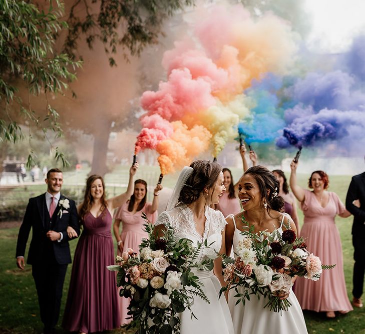 A Lesbian couple celebrate their wedding and are followed by their wedding party who hold colourful smoke bombs in the air.