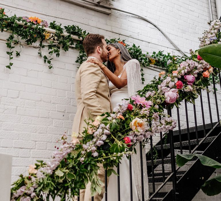 Bride in satin Halfpenny London wedding dress and tasselled bridal cape kisses groom in double breasted linen suit on metal stairs with floral banister decoration at Loft Studios London