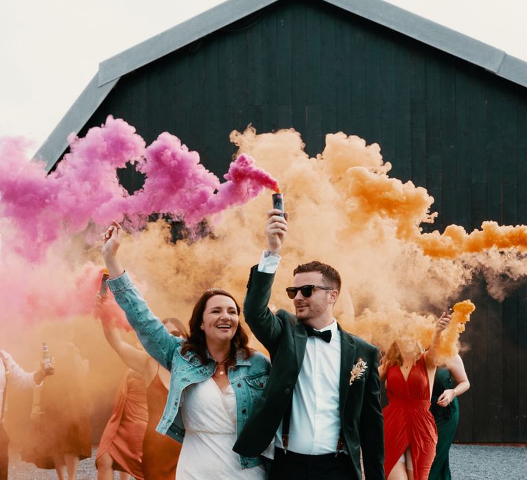 Smoke bomb couples portrait of a bride in a denim jacket holding a pink flare and the groom in black tie suit holding an orange grenade at their autumn wedding 