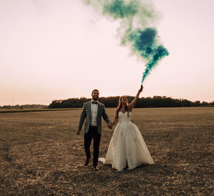 Bride in a Sassi Holford wedding dress holding a green smoke bomb in the air during a golden hour portrait with there husband 