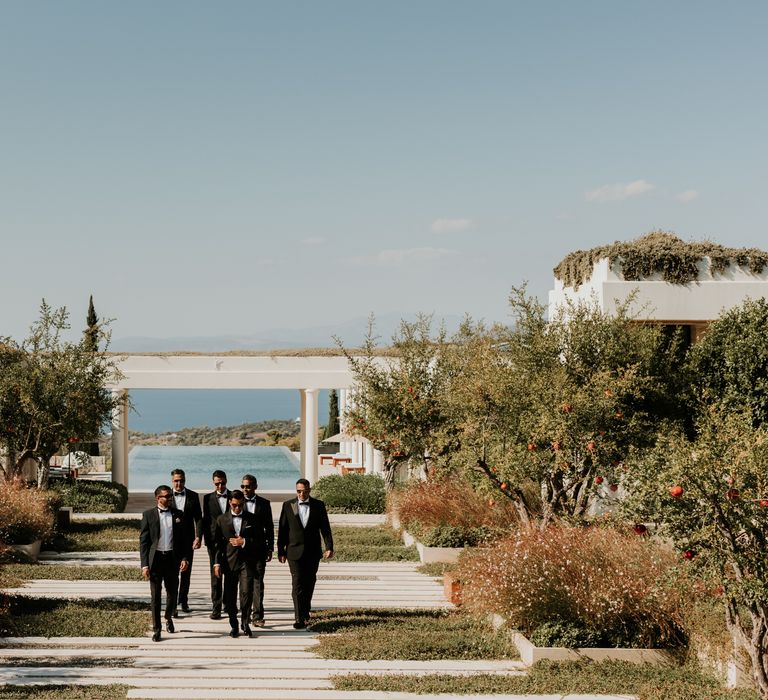 Groom & his groomsmen walk in the distance in front of the sea for Greek wedding | Hannah MacGregor Photo & Film