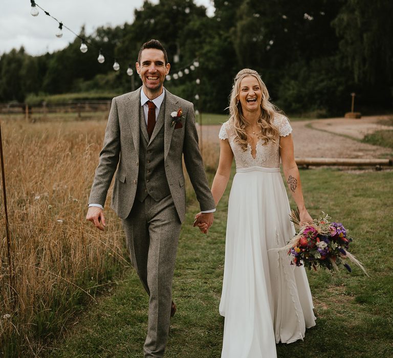 Bride in lace top cap sleeved wedding dress holding mixed bridal bouquet holds hands with groom in grey three piece herringbone suit and red tie as they walk through field at late summer wedding in Norfolk