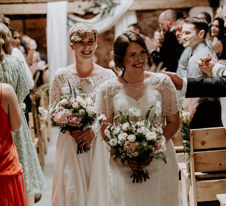 Two brides walking up the aisle holding their pink and white wedding bouquets 