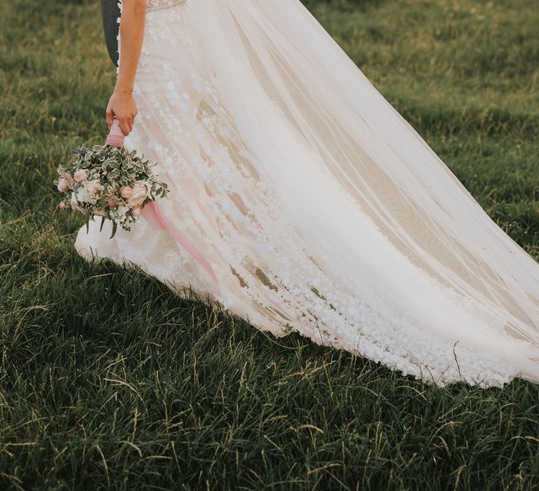 Bride in open back appliqué Berta wedding dress with train and veil holds mixed bridal bouquet as she walks through field with groom during golden hour at Primrose Hill Farm summer wedding