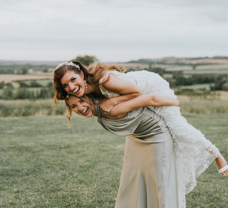 Bride in applique Berta wedding dress and bridal headband laughs on the back of bridesmaid in sage green satin bridesmaid dress in field during summer wedding at Primrose Hill Barn 