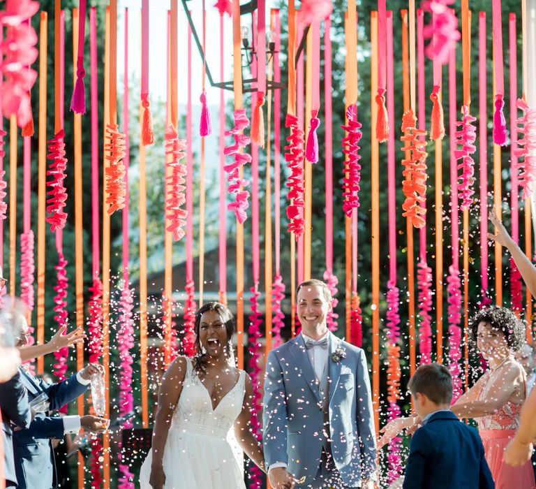 Bride & groom laugh and smile as they walk hand in hand through colourful confetti being thrown by wedding guests