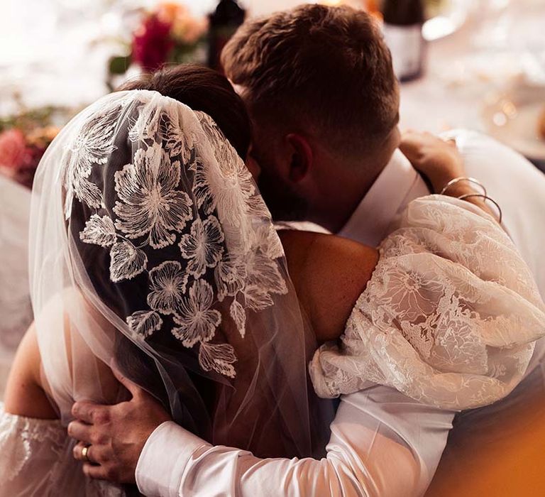 Bride in a lace cap wedding veil with matching detachable puff sleeves embracing her husband at the top table 