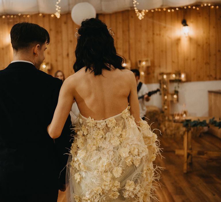 Bride in strapless feathered wedding dress links arm with groom in dark blue suit as they walk down the aisle during rustic wedding ceremony