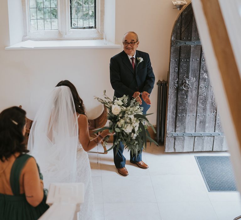 Bride in Pronovias wedding dress and veil carrying green and white rose bridal bouquet walks down stairs with bridesmaids in green bridesmaid dresses towards man in suit with white rose buttonhole before Notley Abbey wedding