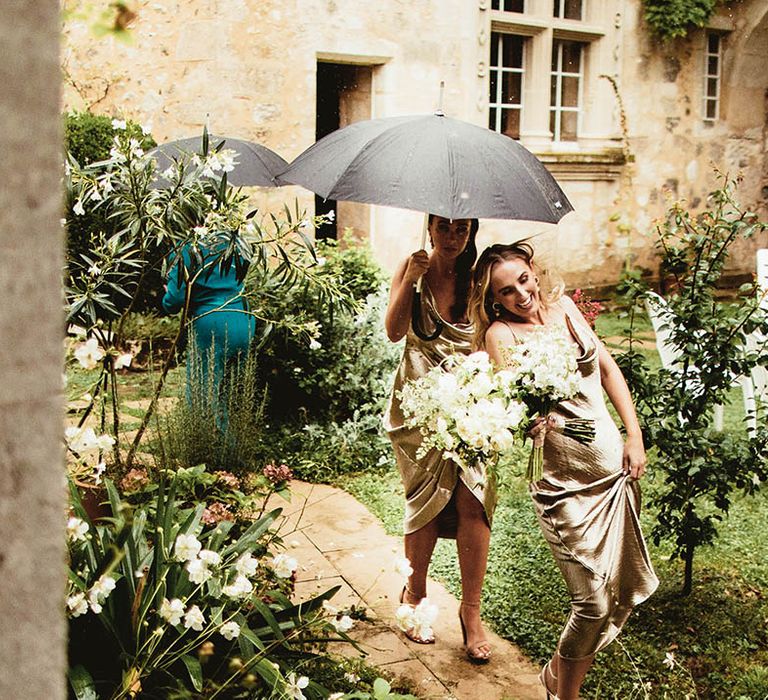 Bridesmaids walk under umbrella on wedding day whilst wearing gold silk dresses