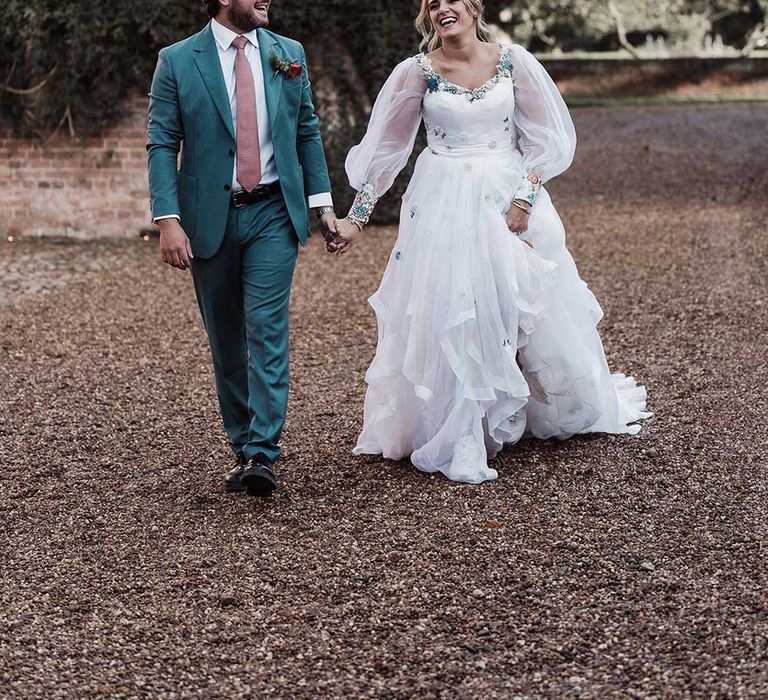 Smiling groom in blue suit and pink tie holding hands with laughing bride in long mesh sleeve wedding dress walks across drive at Iscoyd Park