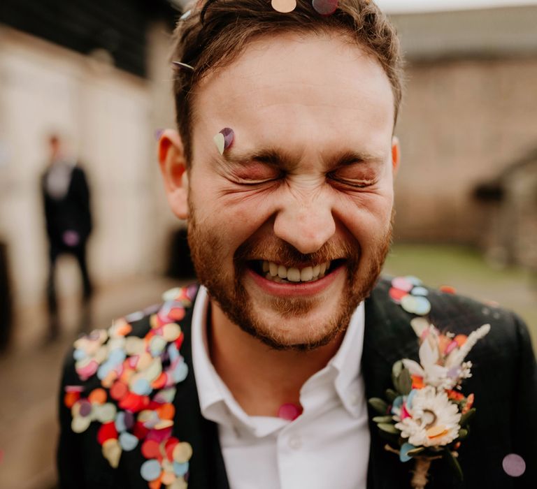 Groom in dark suit and white shirt with floral buttonhole covered in multicoloured confetti smiles at the camera