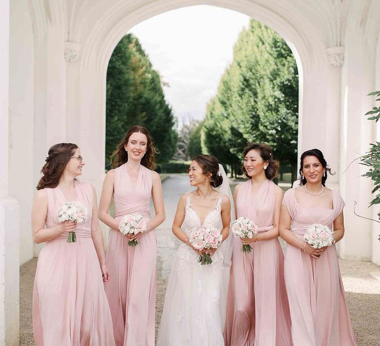 Bride walks with her bridesmaids who wear blush pink gowns with wrapped waistbands and carry white floral bouquets