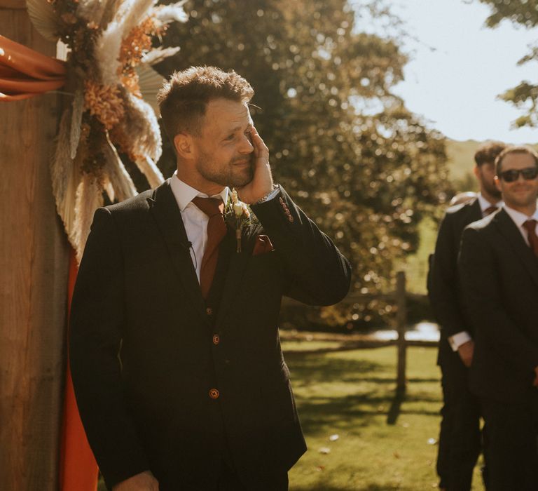 Emotional groom in dark wedding suit and burgundy tie watches bride walk down the aisle during outdoor wedding ceremony